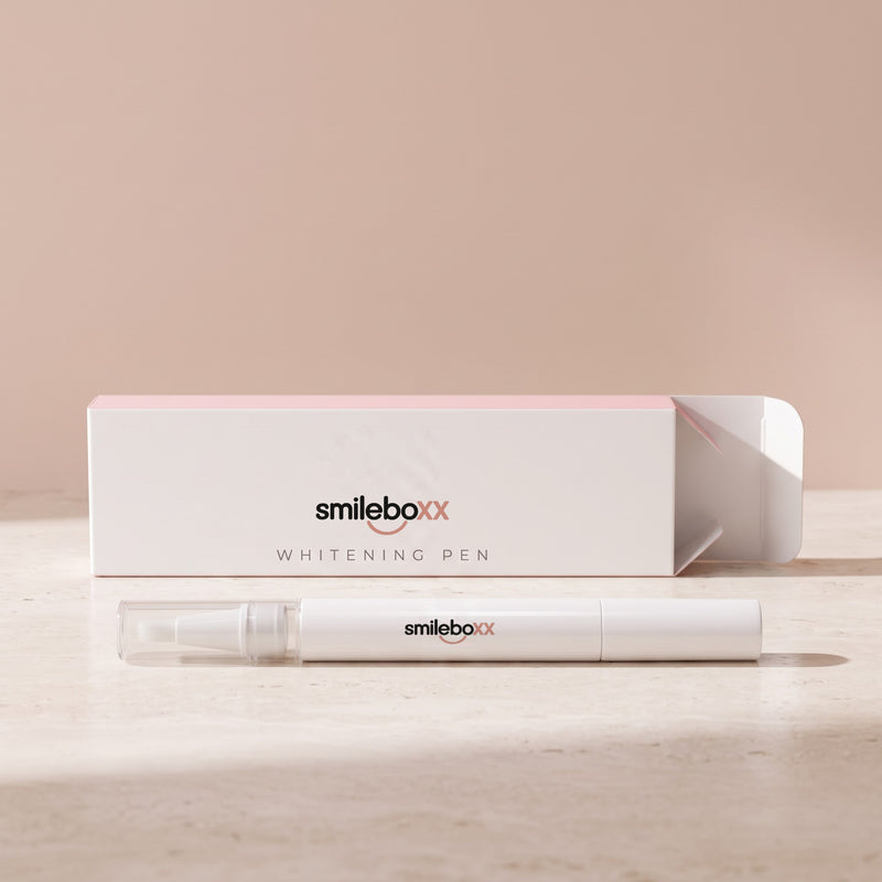A white applicator pen lying in front of an opened carton printed smileboxx and WHITENING PEN