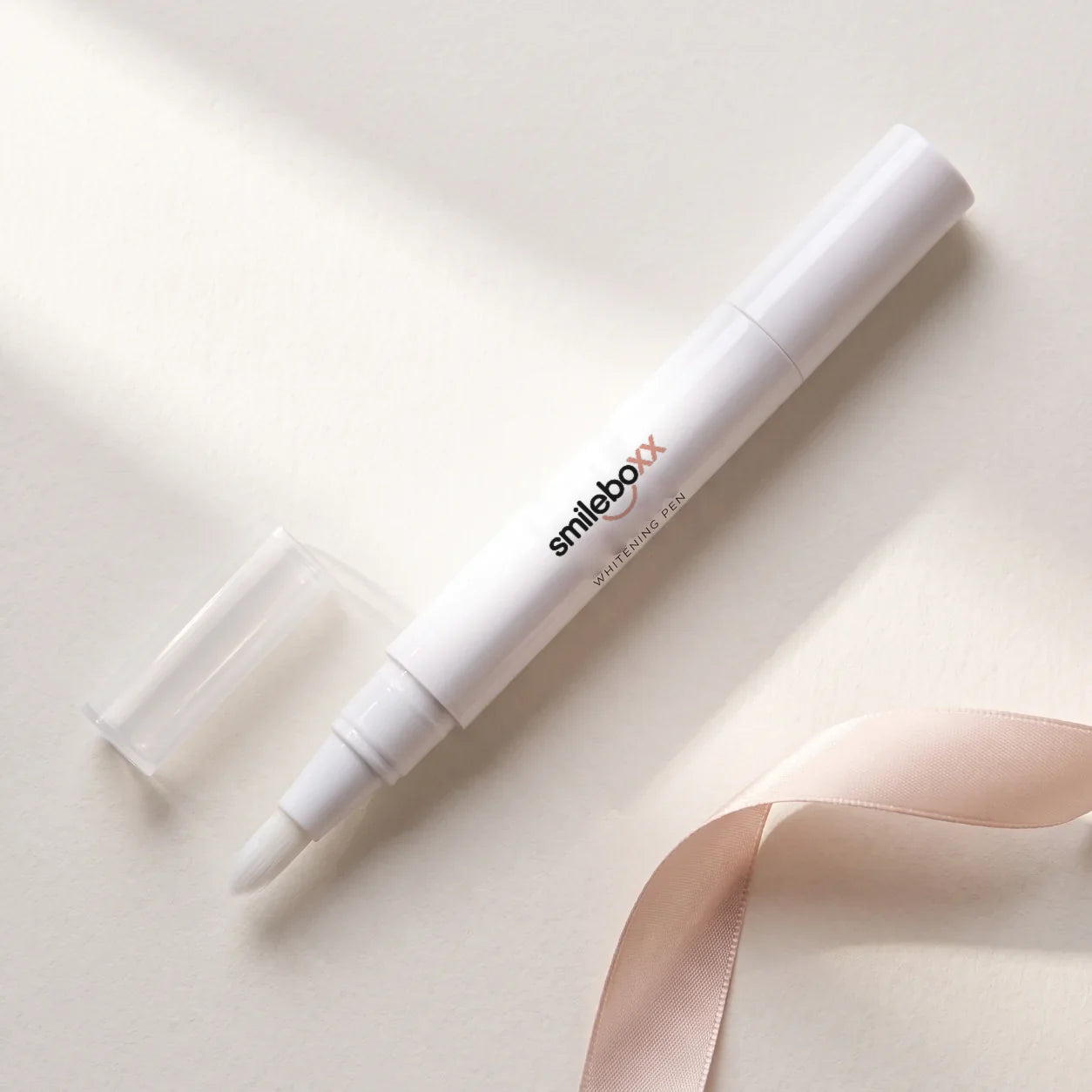 A white applicator pen printed smileboxx and WHITENING PEN lying beside its clear cap and a blush ribbon