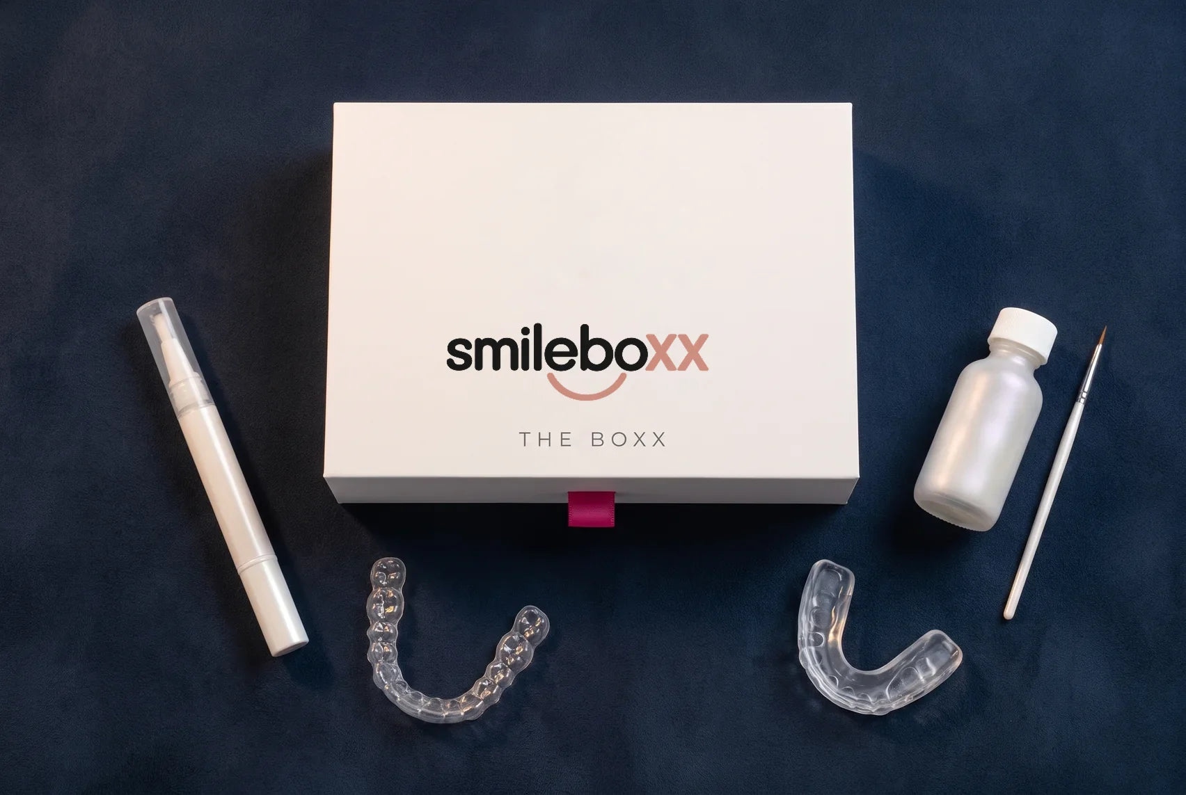 A white box printed smileboxx with a white pen, a clear aligner, a clear guard, a pearl-white bottle and a fine brush on navy suede