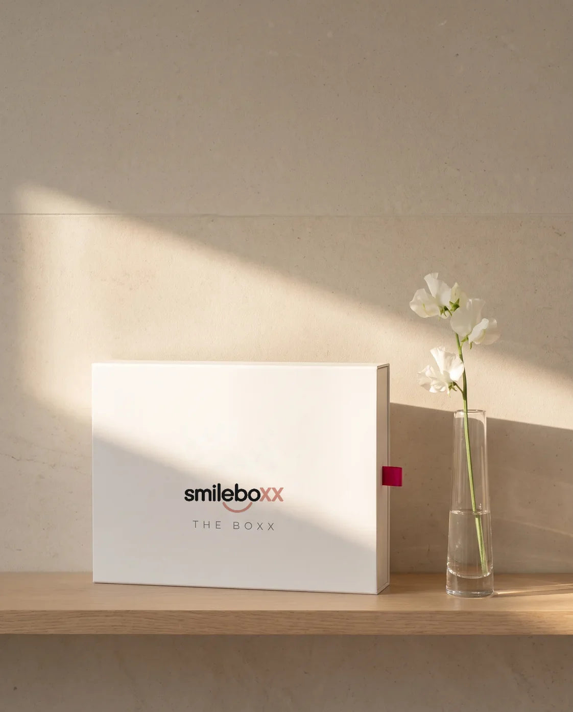 A white box printed smileboxx and THE BOXX standing on an oak shelf beside a sweet pea stem