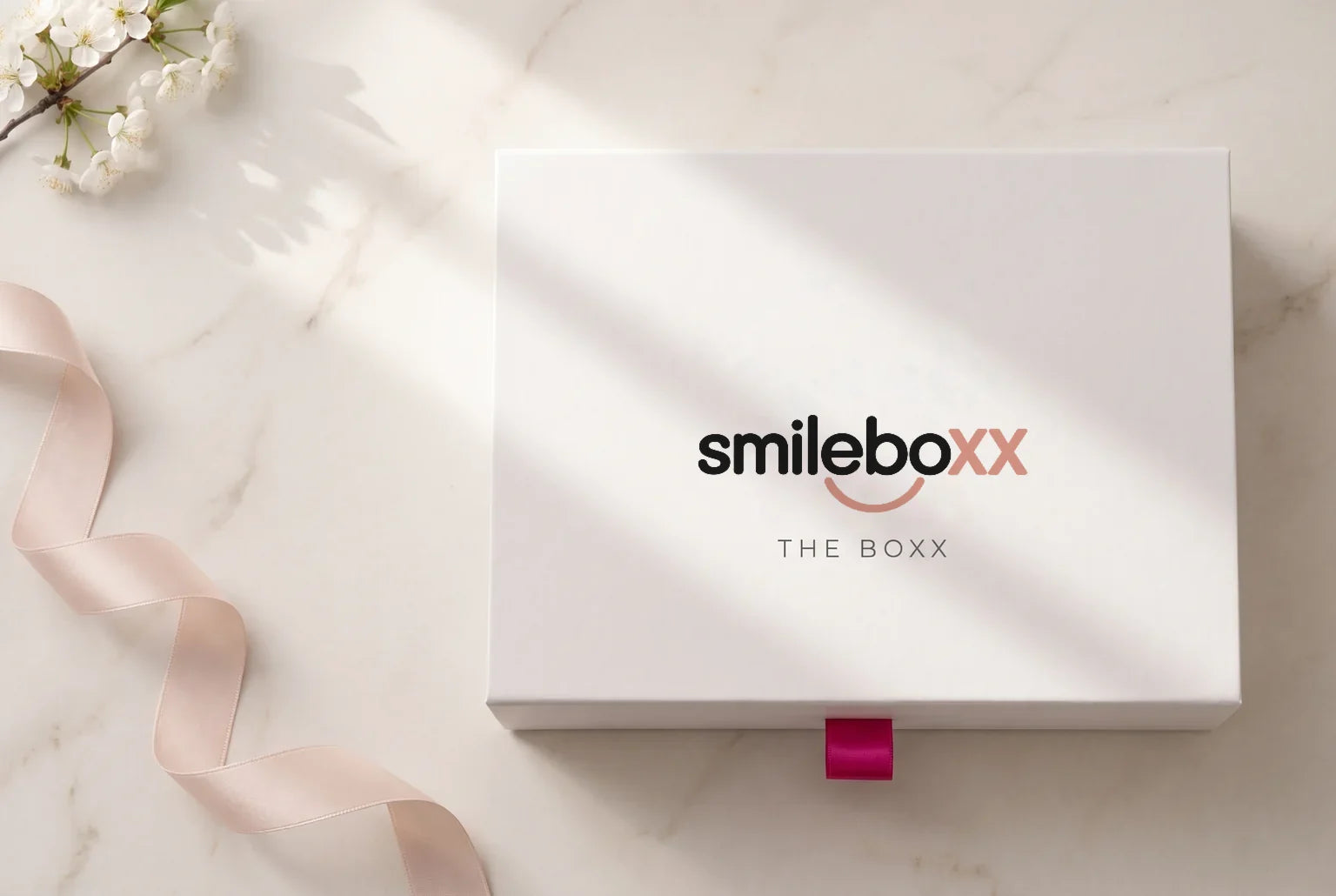 A closed white gift box printed smileboxx and THE BOXX on marble with a blush ribbon and white blossom