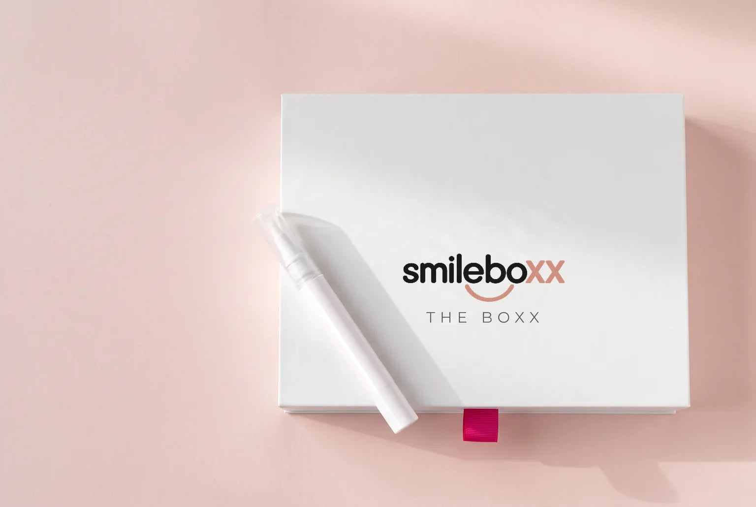A white applicator pen resting on a closed white box printed smileboxx and THE BOXX on blush paper