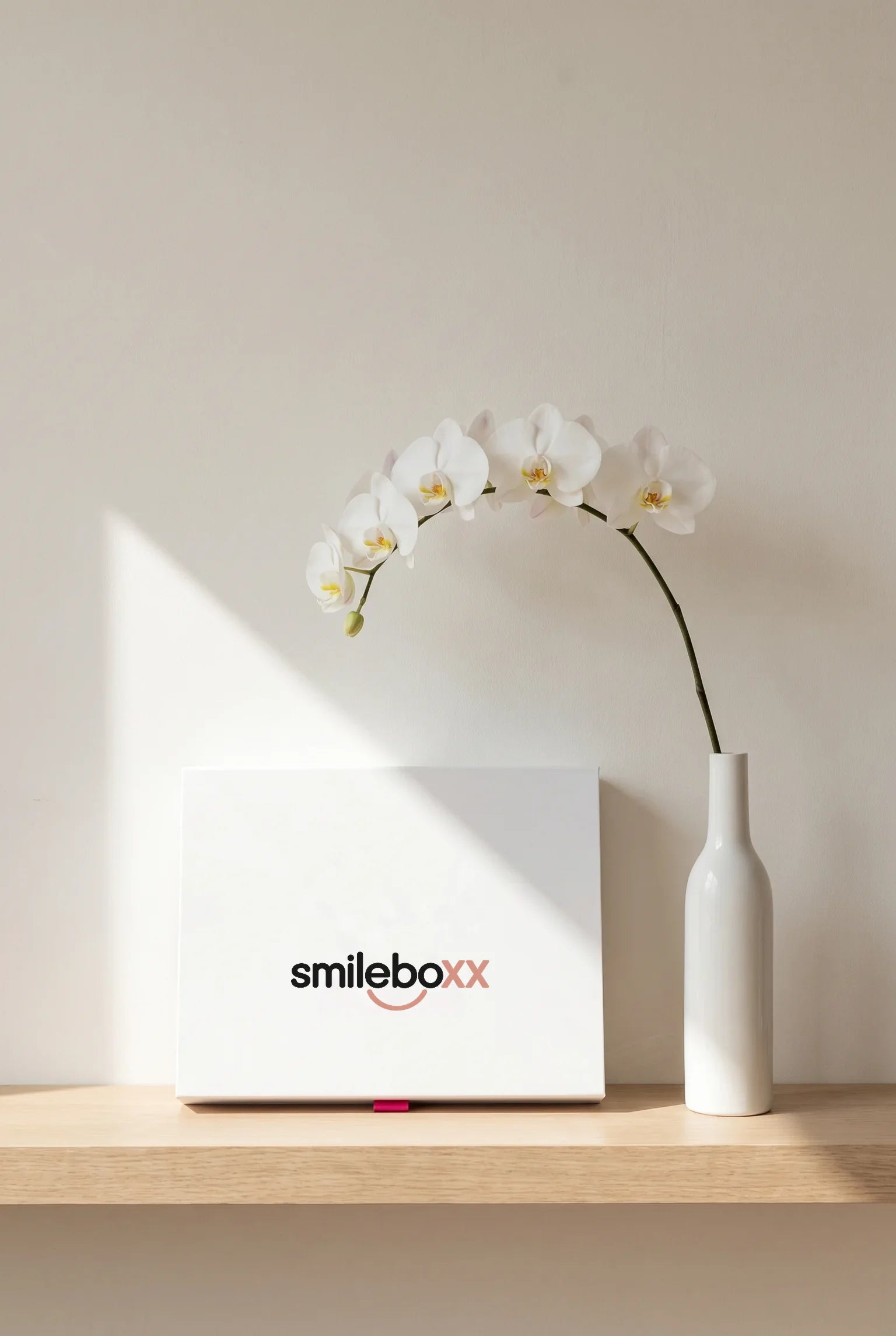 A white box printed smileboxx standing on an oak shelf beside a white orchid in a tall vase