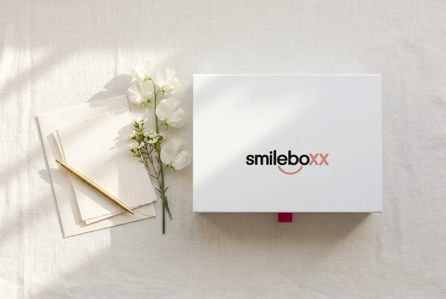 Blank cream notecards, an envelope and a brass pen on a linen desk beside a white box printed smileboxx