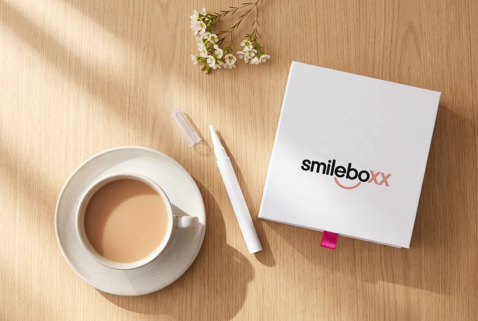 A white box printed smileboxx on an oak table with a cup of tea, a white applicator pen and white waxflower